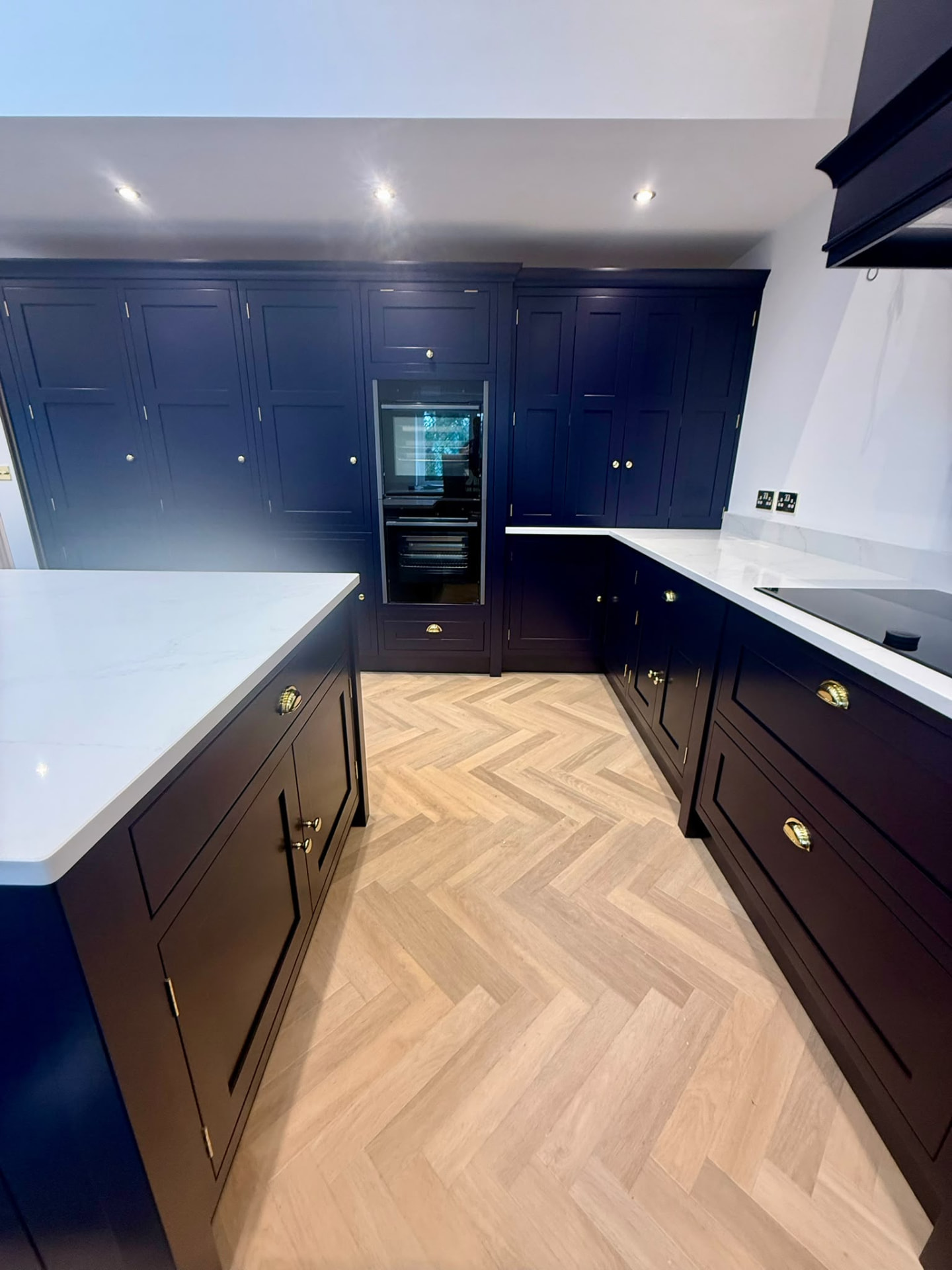 Full view of bespoke navy hand painted kitchen with island, brass hardware and pale herringbone floor