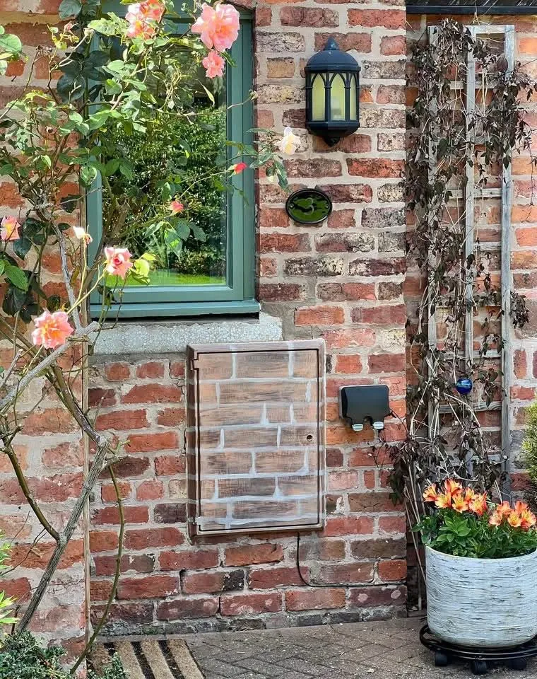 Sage green painted window frame and hand-painted brick-effect utility cover on a brick garden elevation
